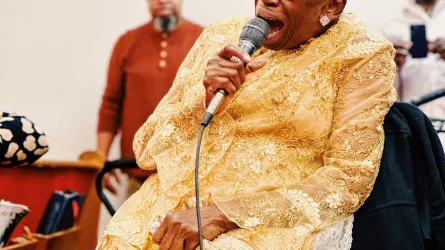 Mother Perry singing into a handheld microphone, wearing a gold long-sleeve dress.